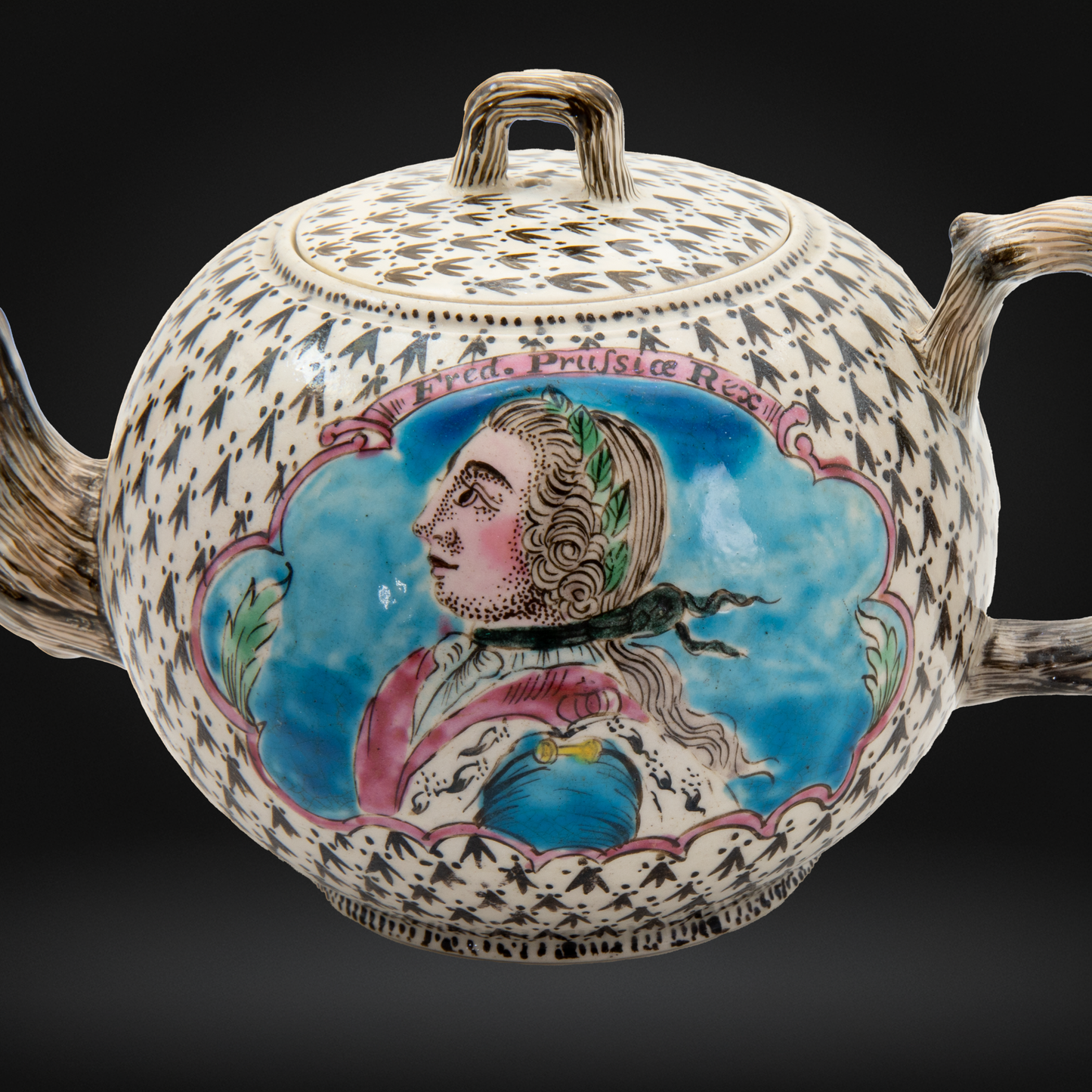An antique porcelain teapot with a detailed hand-painted portrait of the King of Prussian, King Frederick II. on the front, set against an urbine background.