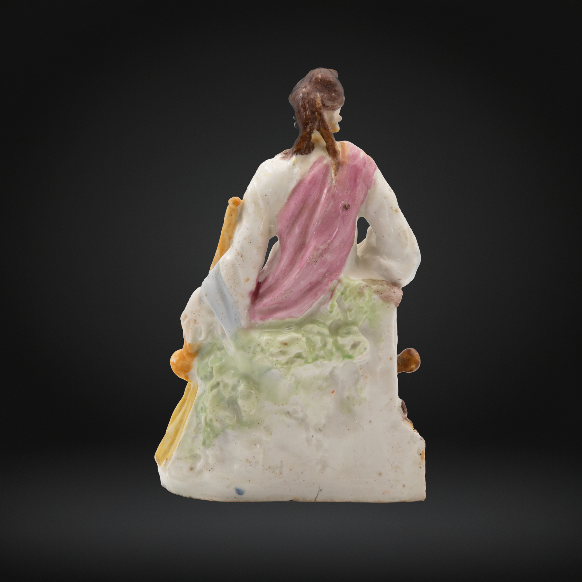 A hand-painted porcelain figure of a seated muse Euterpe viewed from behind, featuring a white base with a pink draped shawl, green patterned skirt details, and brown hair.